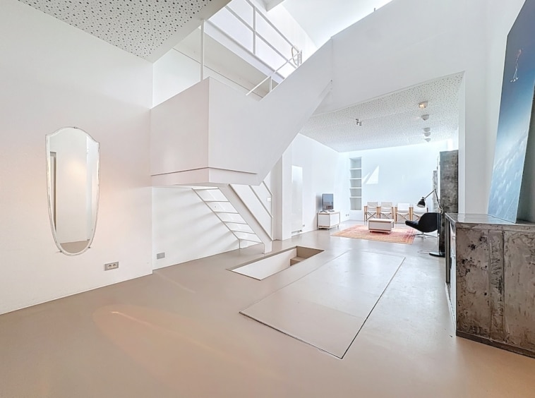 Salon moderne lumineux avec escalier angulaire, murs blancs, béton industriel et décoration minimaliste.