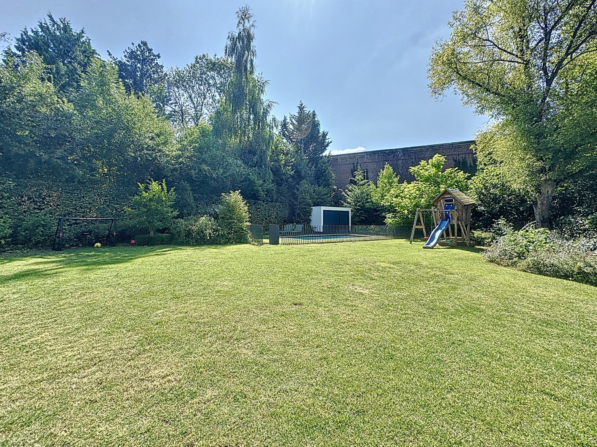 Jardin en famille ensoleillé avec pelouse, aire de jeux en bois, arbres et nature luxuriante