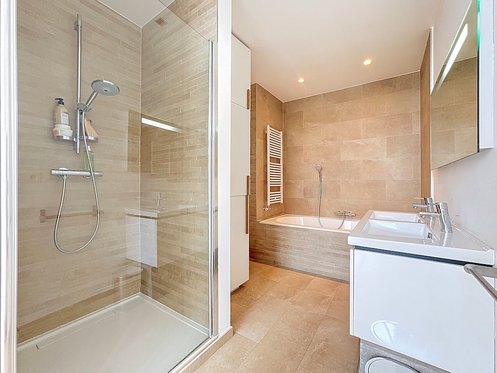 Salle de bain moderne avec douche en verre, baignoire élégante et double lavabo lumineux.
