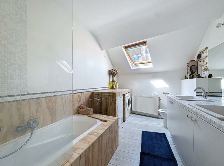 Salle de bain moderne en attique avec baignoire, douche en verre, lumière naturelle et design épuré.