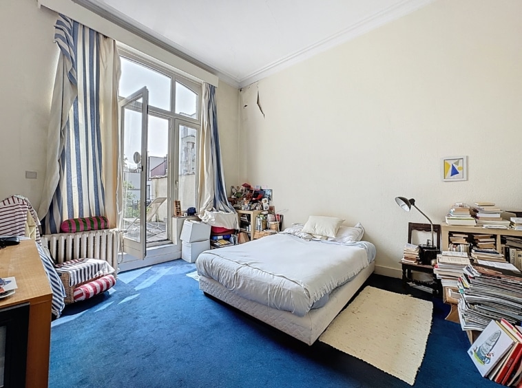 Chambre lumineuse avec lit gris, balcon ensoleillé, bureau en bois, décoration artistique et apaisante.