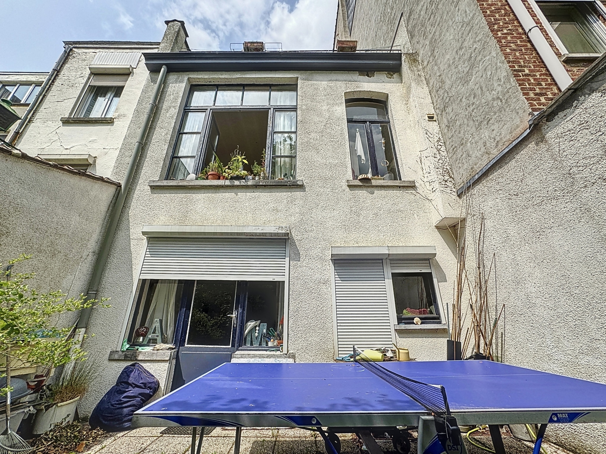 Maison lumineuse avec terrasse et tables de ping-pong, entourée d’un jardin verdoyant.