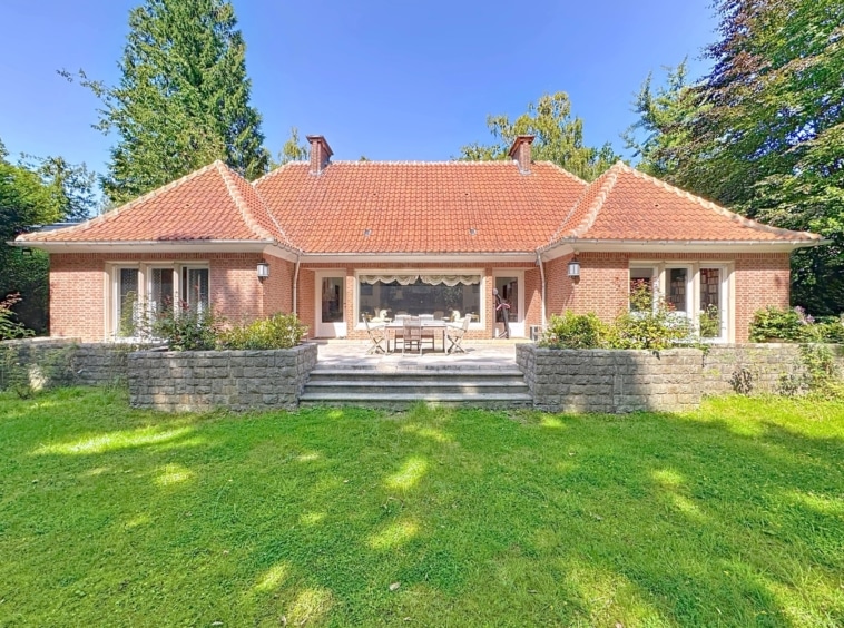 Charmante maison en brique avec terrasse, jardin verdoyant et vue sur grands arbres sous ciel bleu.