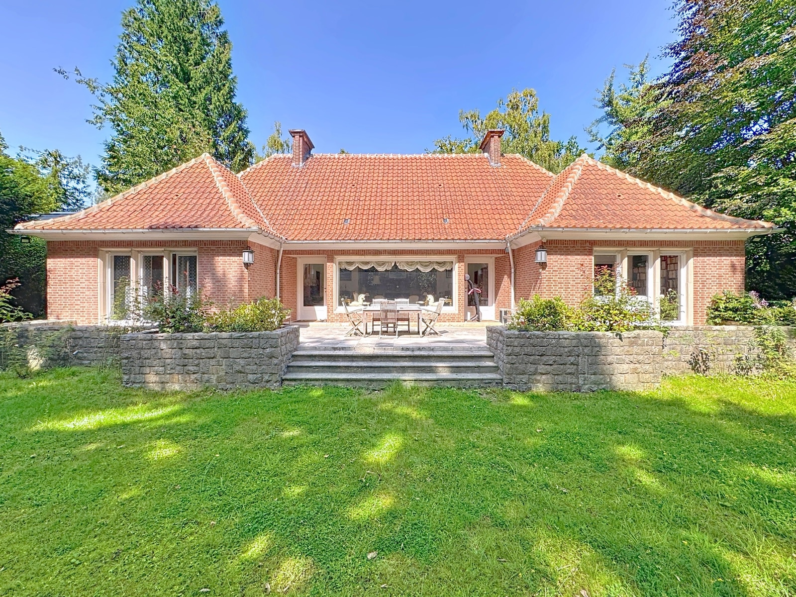 Charmante maison en brique avec terrasse, jardin verdoyant et vue sur grands arbres sous ciel bleu.