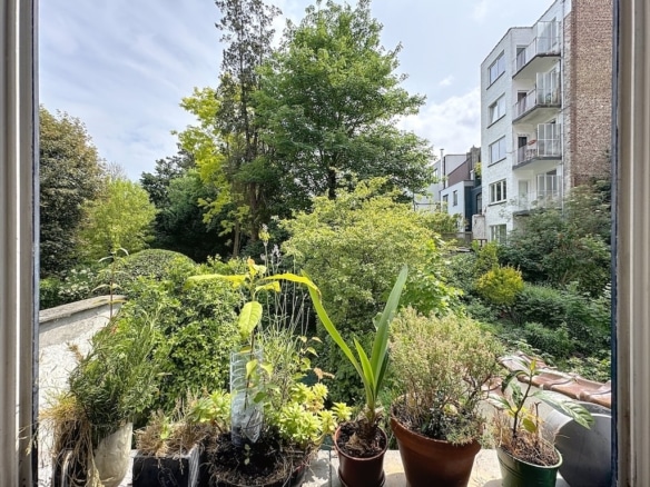 Balcon végétalisé offrant vue sur jardin urbain, arbres, buissons, immeubles modernes et ciel partiellement nuageux.