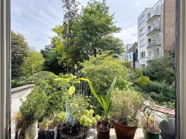 Balcon végétalisé offrant vue sur jardin urbain, arbres, buissons, immeubles modernes et ciel partiellement nuageux.