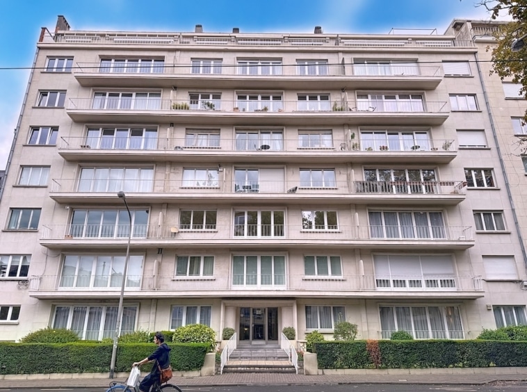 Appartement moderne avec balcons, façade en béton clair, jardin urbain et street view.