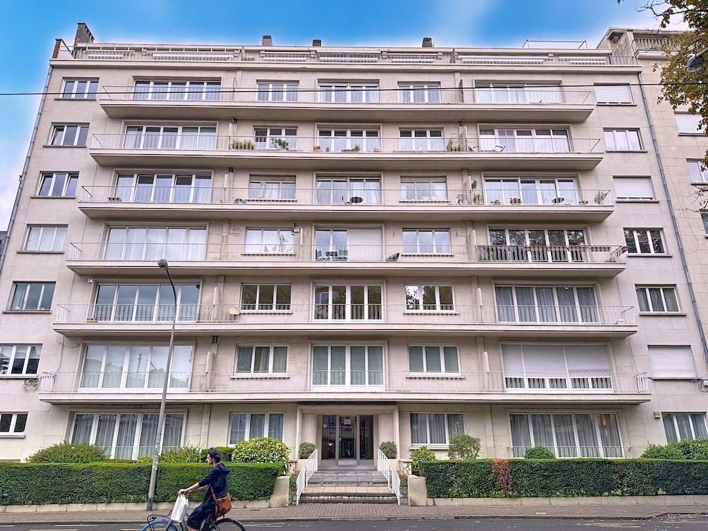 Appartement moderne avec balcons, façade en béton clair, jardin urbain et street view.