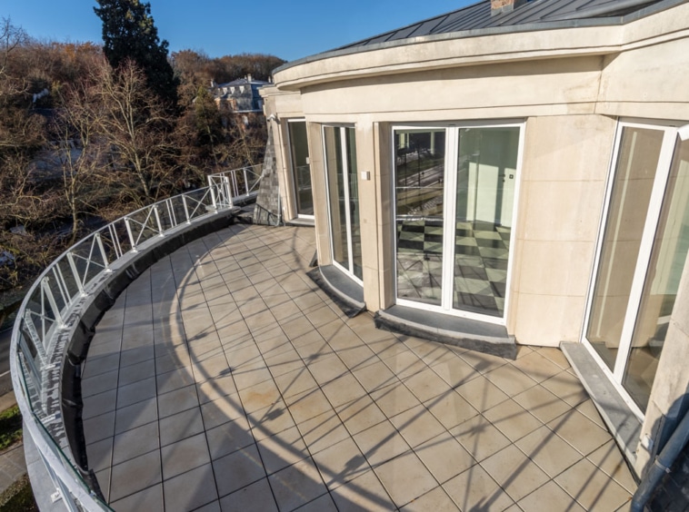 Balcon courbé moderne avec vue panoramique, verre, bois et verdure, idéal pour détente et apéritifs.