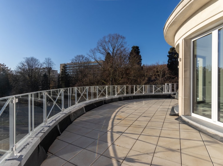 Balcon moderne courbé avec vue sur nature, terrasse en tuiles, rambarde en verre, extérieur lumineux.