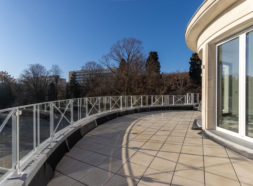 Balcon moderne courbé avec vue sur nature, terrasse en tuiles, rambarde en verre, extérieur lumineux.