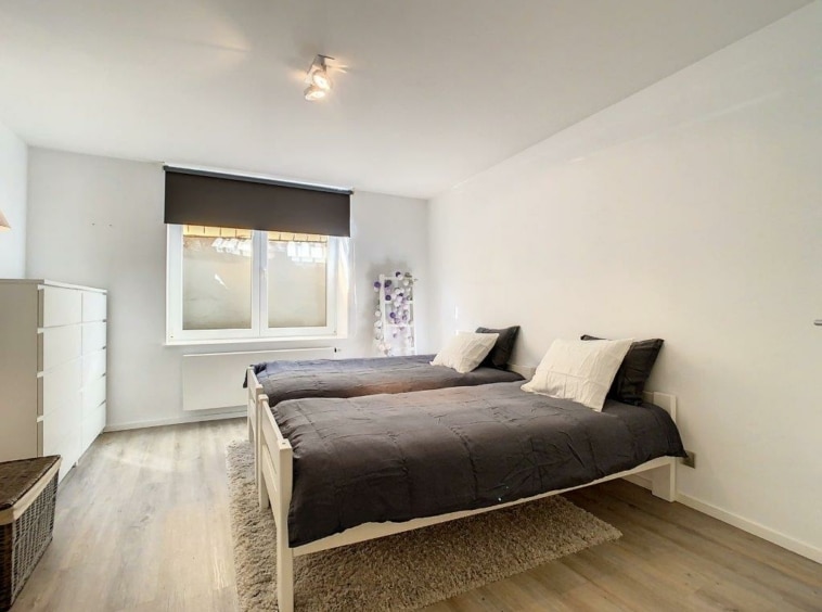 Chambre minimaliste lumineuse avec lits gris, grande fenêtre et rangements modernes.