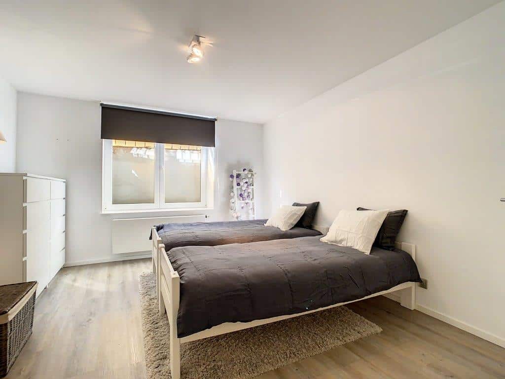 Chambre minimaliste lumineuse avec lits gris, grande fenêtre et rangements modernes.