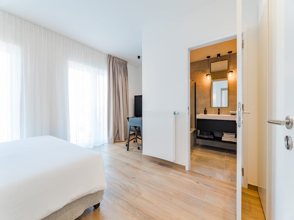 Chambre moderne avec lit blanc, espace de travail, lumière naturelle et salle de bain attenante.