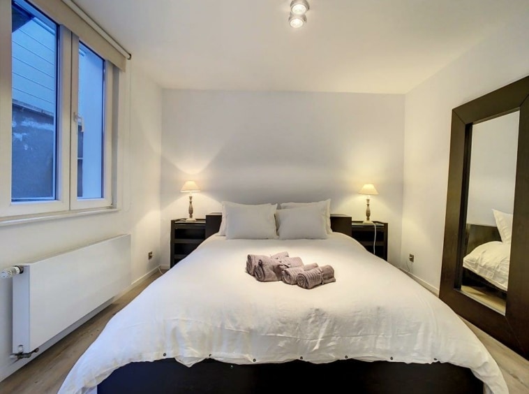 Chambre moderne lumineuse avec grand lit blanc, miroir élégant, lampes douces et sol en bois chaleureux.