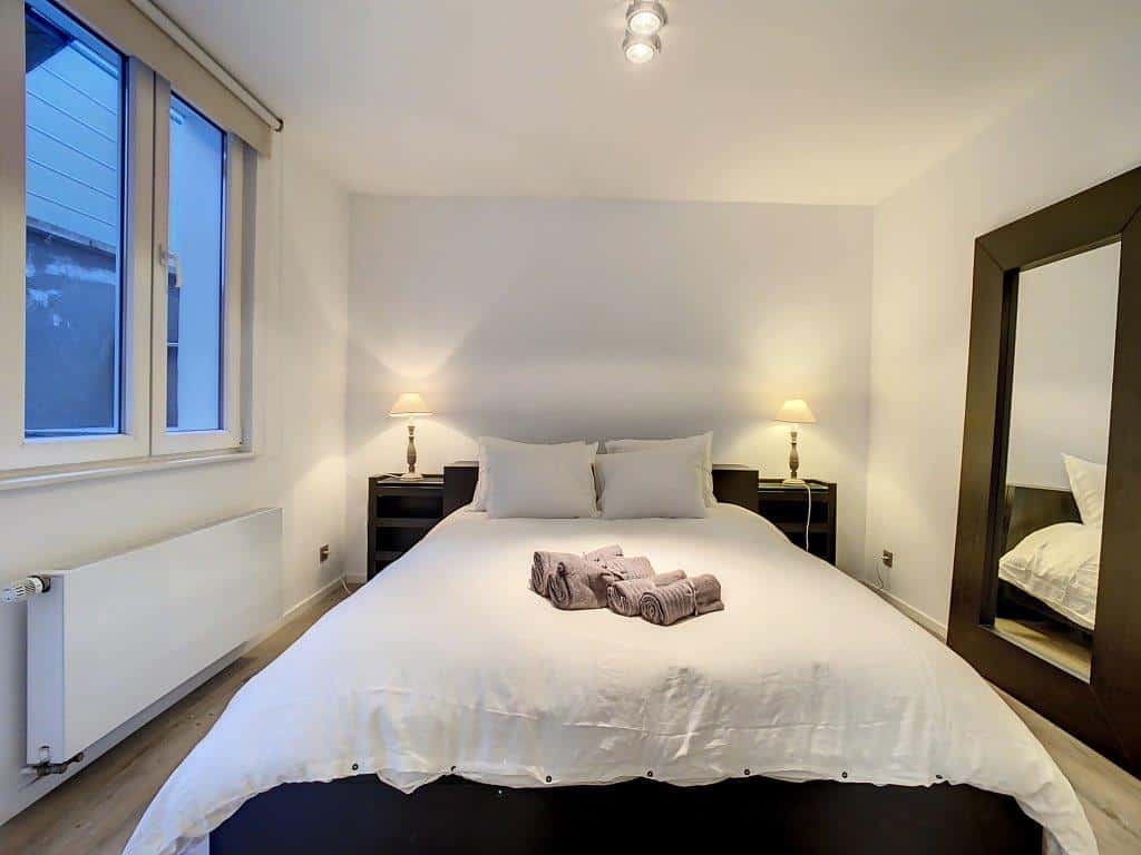 Chambre moderne lumineuse avec grand lit blanc, miroir élégant, lampes douces et sol en bois chaleureux.