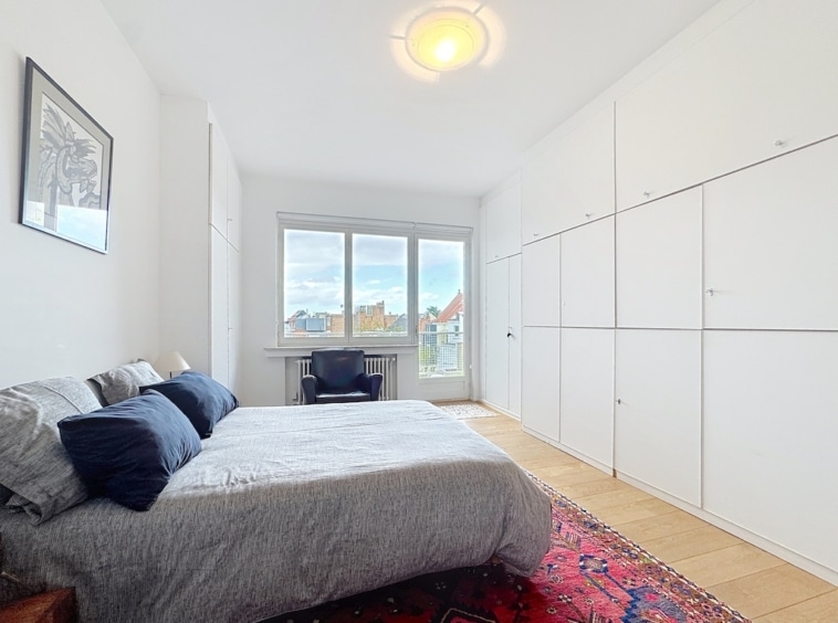 Chambre minimaliste moderne avec vue urbaine lumineuse, lits gris, mobilier blanc intégré et espace de travail.