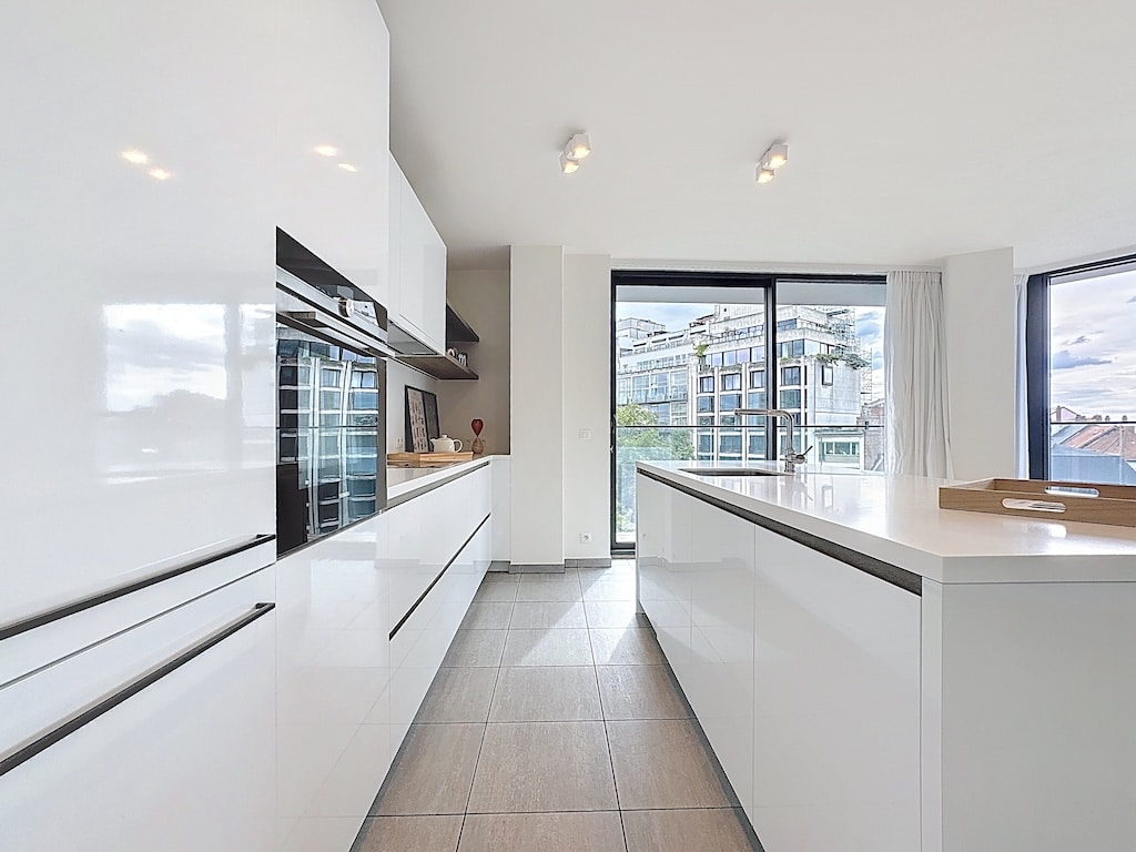 Cuisine moderne blanche lumineuse avec îlot central, grandes fenêtres et appareils intégrés.