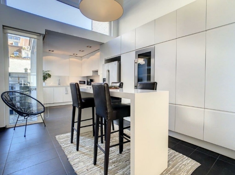 Cuisine moderne minimaliste avec îlot blanc, vue extérieure, mobilier élégant et ambiance lumineuse.