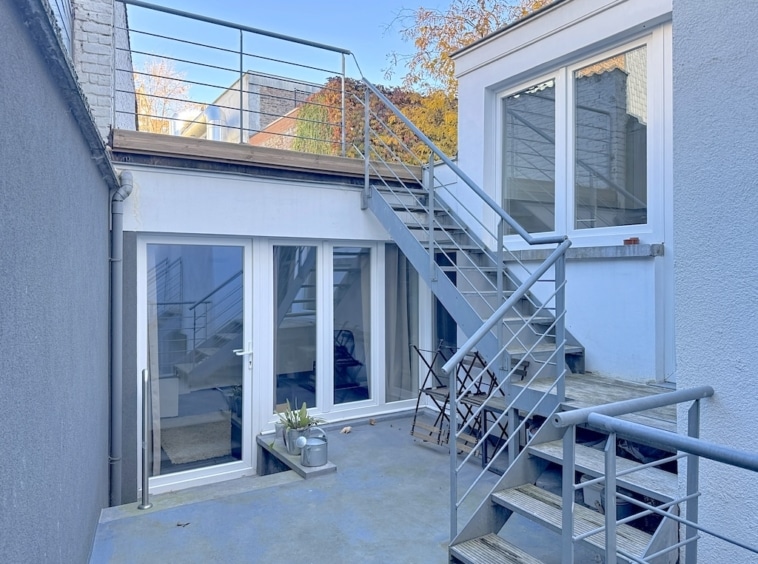 Espace extérieur moderne avec escalier en métal, murs blancs, plantes, vue urbaine et lumière naturelle.