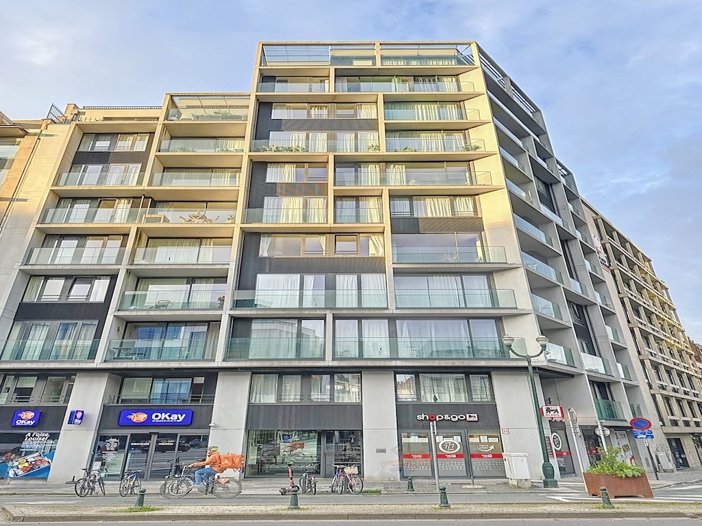 Immeuble moderne en verre avec balcons, commerces, espace piéton végétalisé en centre urbain.