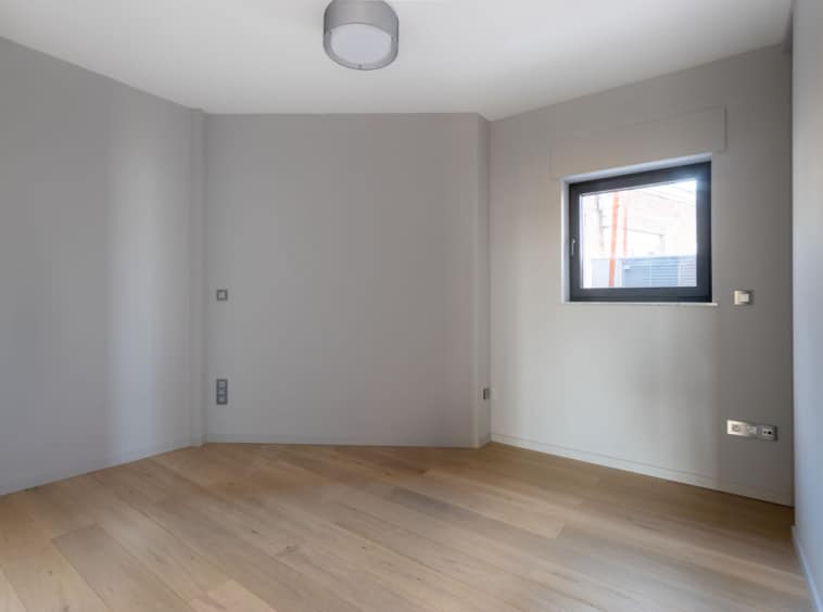 Espace minimaliste lumineux en gris clair avec grande fenêtre, sol en bois et éclairage moderne.