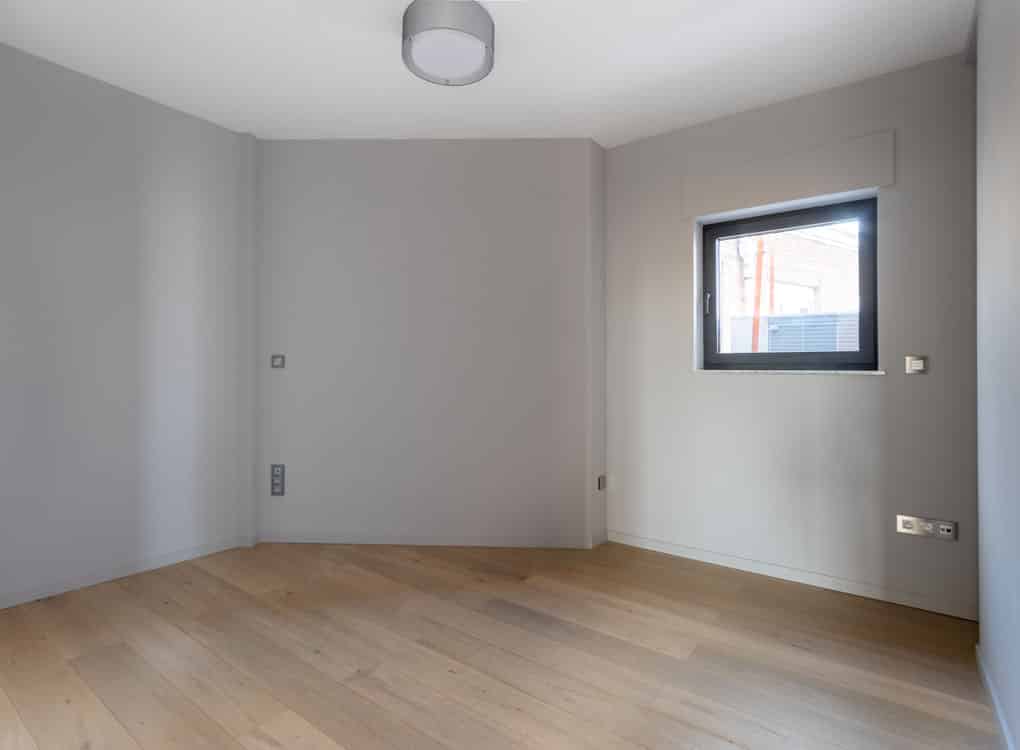Espace minimaliste lumineux en gris clair avec grande fenêtre, sol en bois et éclairage moderne.