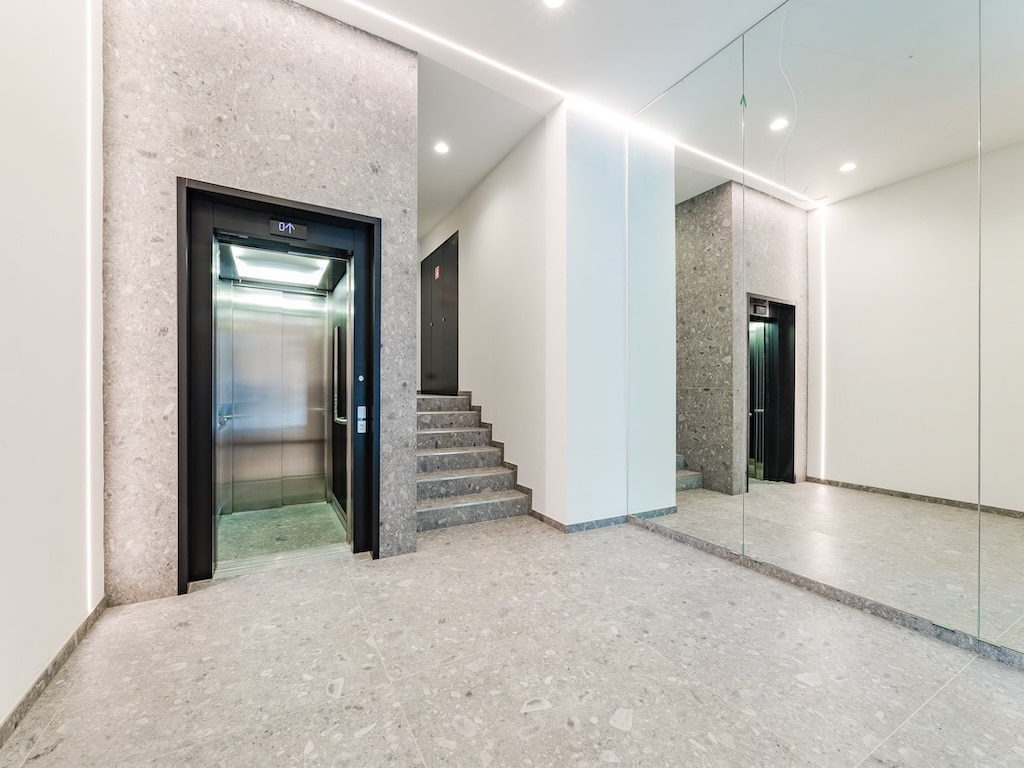Hall d’ascenseur moderne avec miroir, escalier contemporain et éclairage élégant.