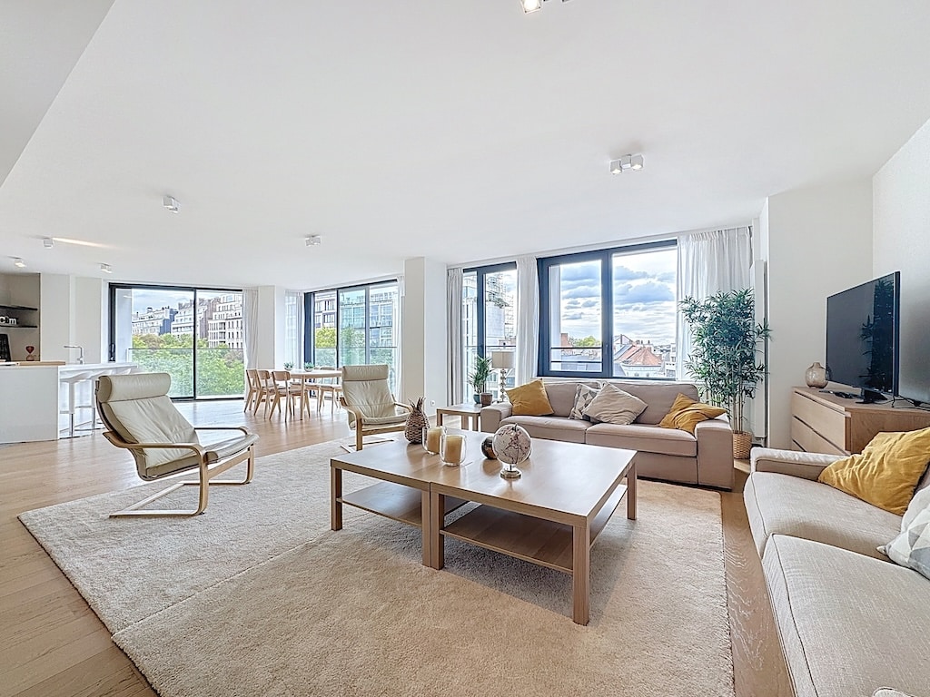 Salon moderne lumineux avec grandes fenêtres, mobilier en bois clair, vue extérieure et ambiance chaleureuse.