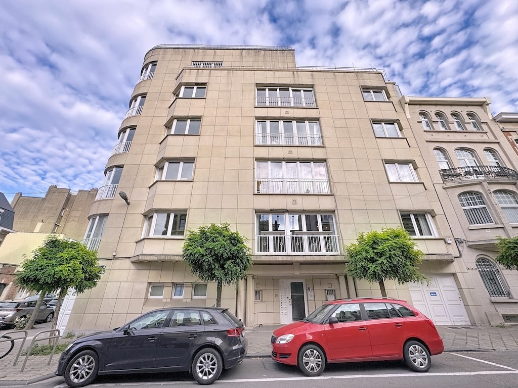 Bâtiment moderne avec façade courbée, grandes fenêtres, parking, arbres et ciel bleu nuagé.