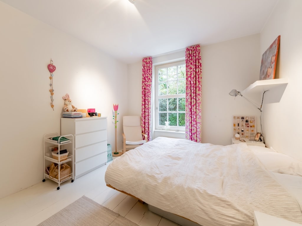 Chambre lumineuse avec lit crème, rideaux roses, mobilier blanc et ambiance chaleureuse.
