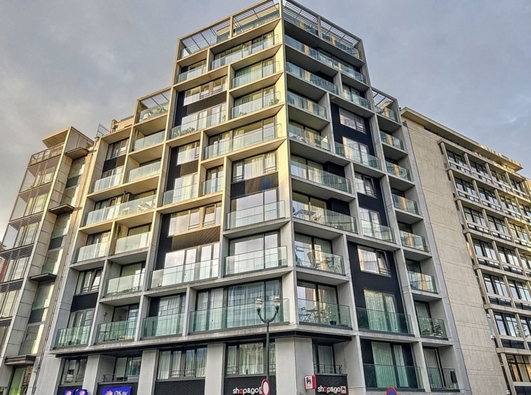 Immeuble contemporain avec façades en verre, balcons verdoyés, et commerces en rez-de-chaussée.