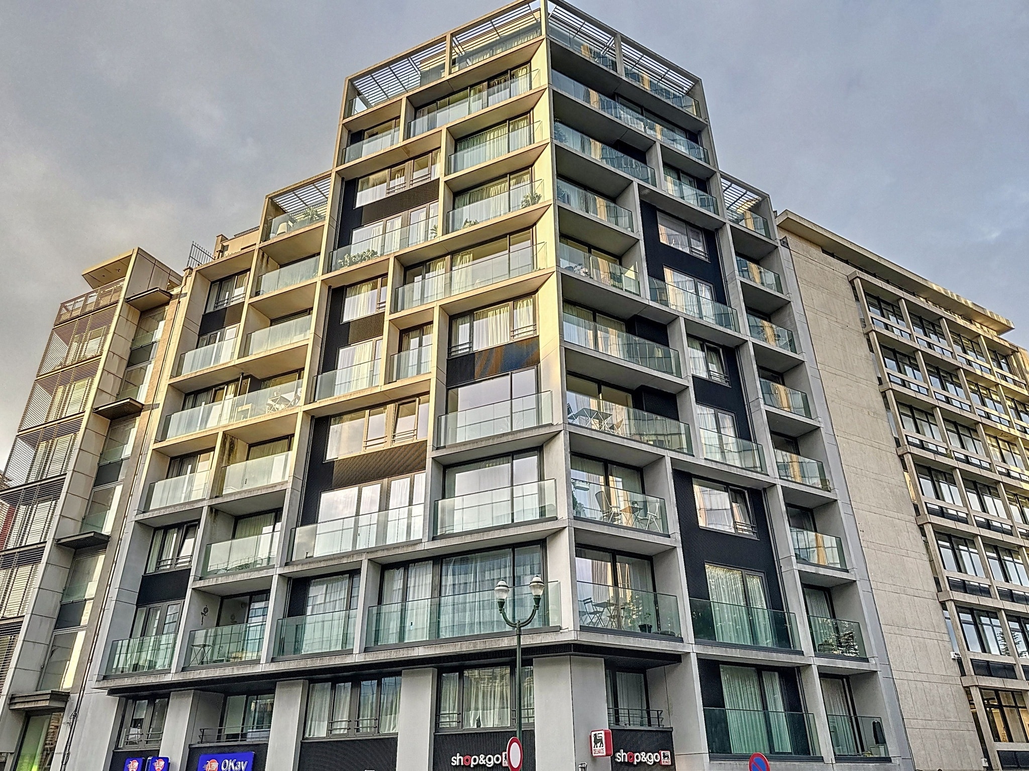 Immeuble contemporain avec façades en verre, balcons verdoyés, et commerces en rez-de-chaussée.