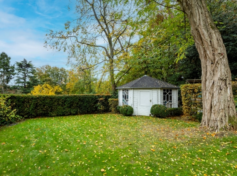 Jardin automnal avec cabane blanche, haies bien taillées et arbres dorés ensoleillés.