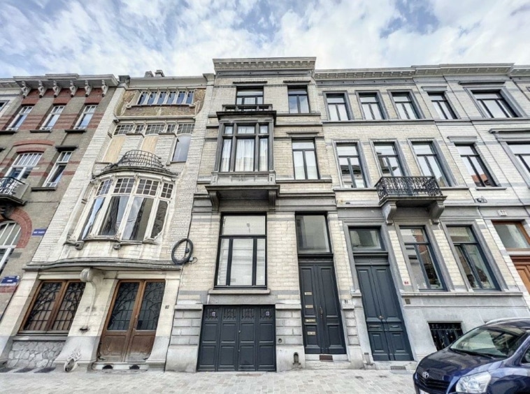 Rue pittoresque avec bâtiments en pierre, balcons, façades élégantes et ciel nuageux.