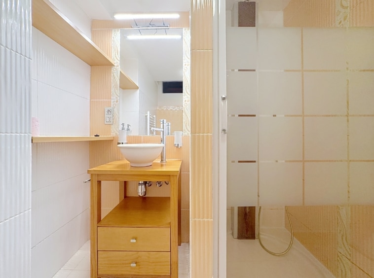 Salle de bain moderne, lumineuse, épurée avec lavabo, rangement en bois et cabine de douche en verre.