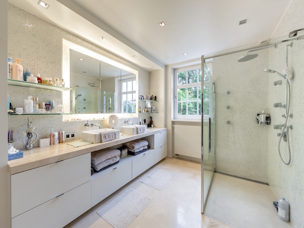 Salle de bain moderne lumineuse avec double vasque, douche à litalienne et vue verdoyante.