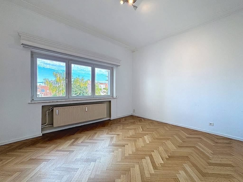 Salle lumineuse avec parquet en chevron, murs blancs et vue verdoyante.