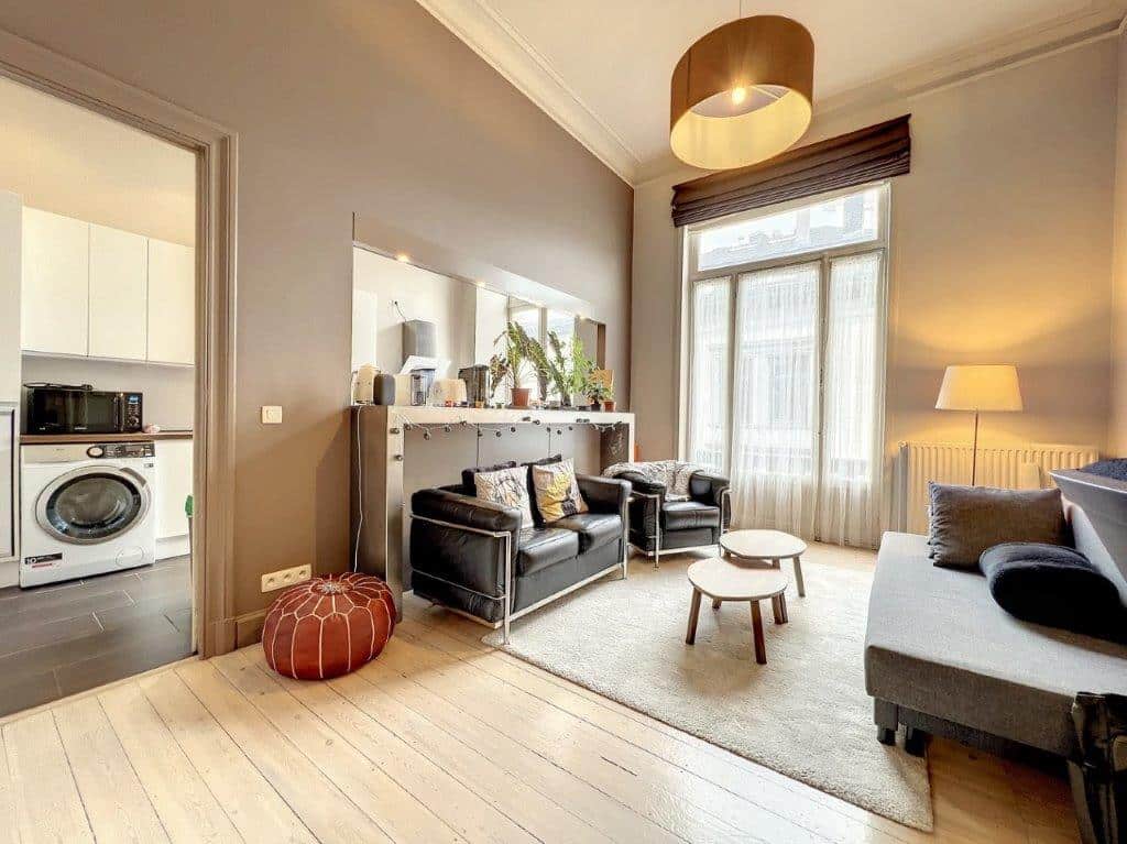 Salon moderne lumineux avec canapé en cuir, plantes, miroirs et cuisine ouverte élégante.