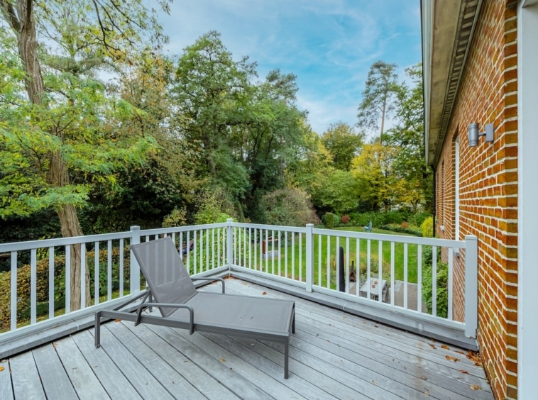 Terrasse en bois avec fauteuil gris, vue automnale sur jardin verdoyant et ciel bleu doux.