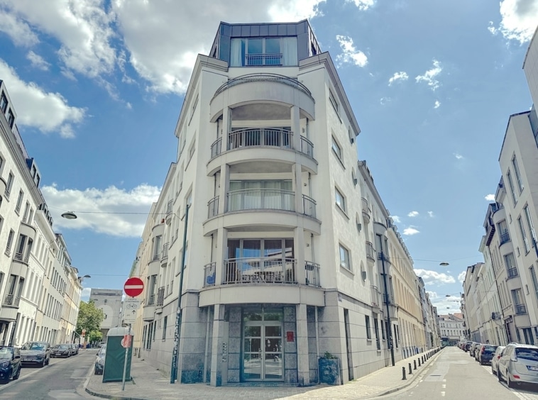 Immeuble moderne blanc aux balcons arrondis, au croisement de rues, sous un ciel clair et nuageux.