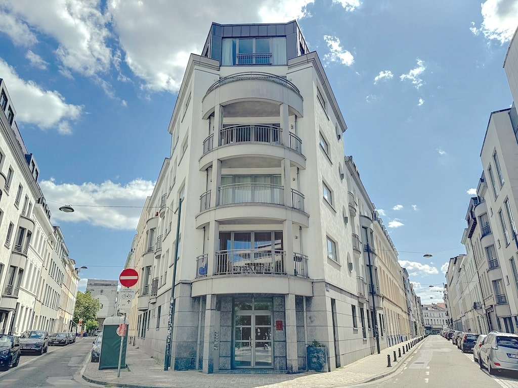 Immeuble moderne blanc aux balcons arrondis, au croisement de rues, sous un ciel clair et nuageux.