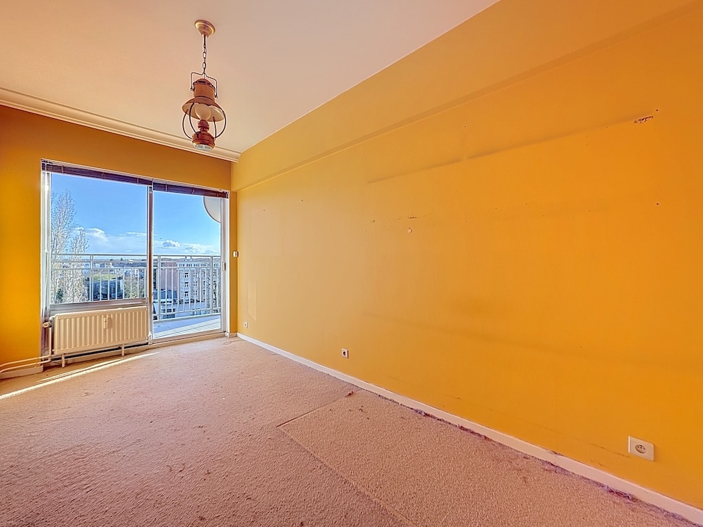Chambre lumineuse aux murs jaunes, grande fenêtre, volets blancs, plafond avec ventilateur rustique.