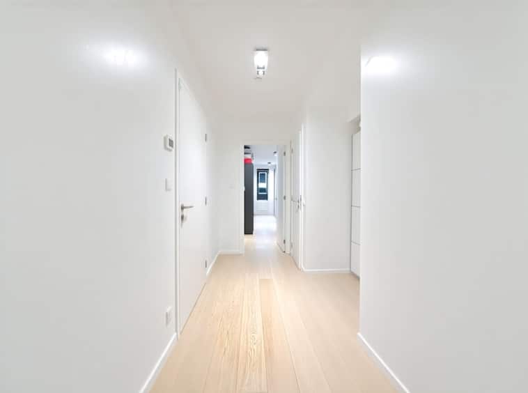 Couloir moderne lumineux avec murs blancs, sol en bois clair, portes discrètes et ambiance épurée.