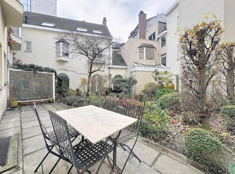 Jardin urbain paisible avec table en pierre, chaises noires, végétation, arbre naissant, sous ciel nuageux.
