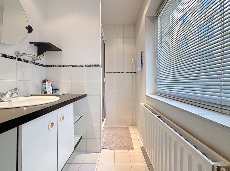 Salle de bain moderne avec douche en verre, carrelage blanc, meubles noirs et grande fenêtre lumineuse.
