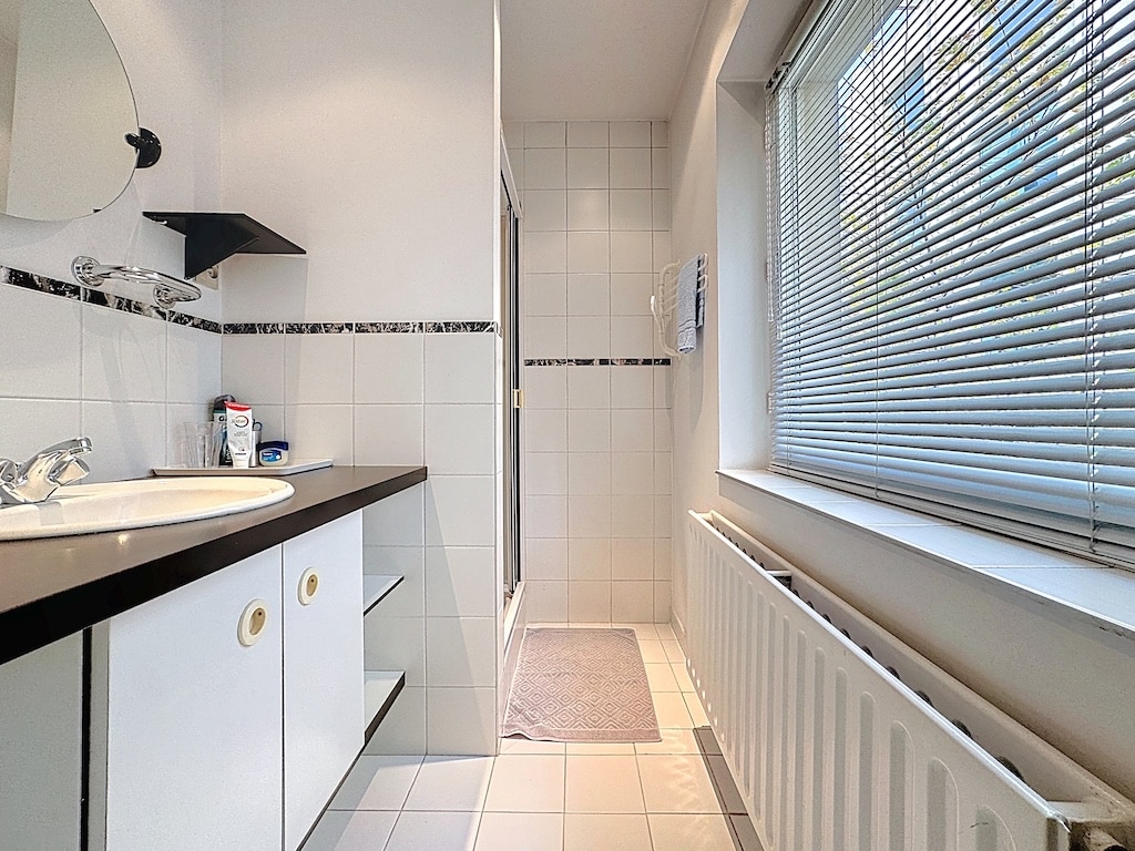 Salle de bain moderne avec douche en verre, carrelage blanc, meubles noirs et grande fenêtre lumineuse.
