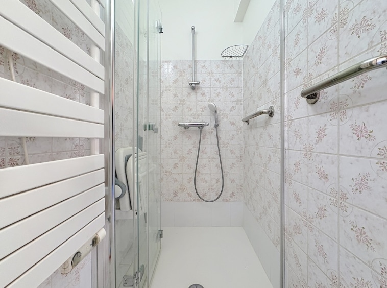 Salle de douche moderne avec carrelage floral, douche à pluie, porte-serviettes chauffant et barres de maintien.