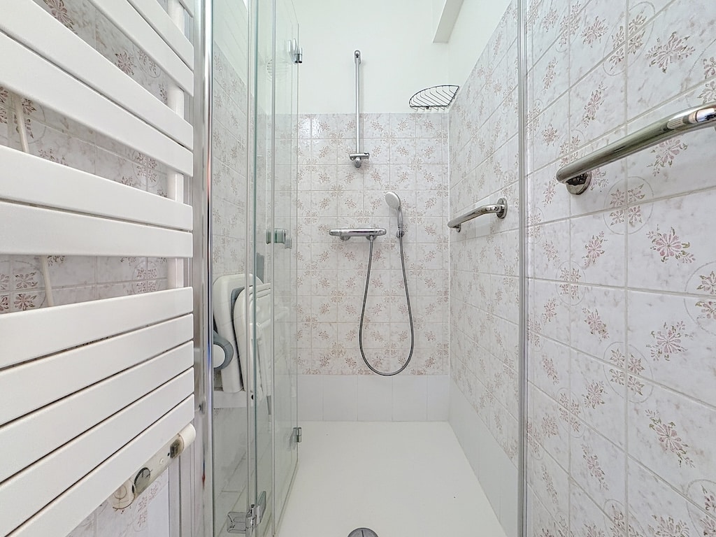 Salle de douche moderne avec carrelage floral, douche à pluie, porte-serviettes chauffant et barres de maintien.
