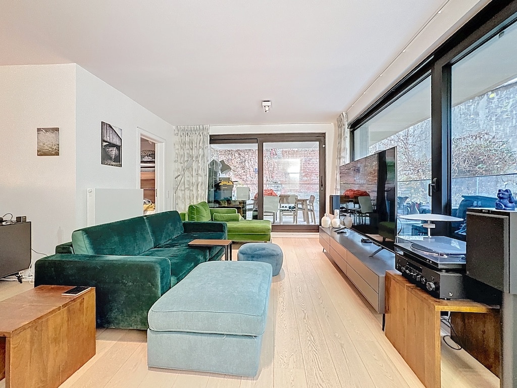 Salon moderne lumineux avec canapé velours vert, ottoman bleu, grand fenêtres et espace convivial.